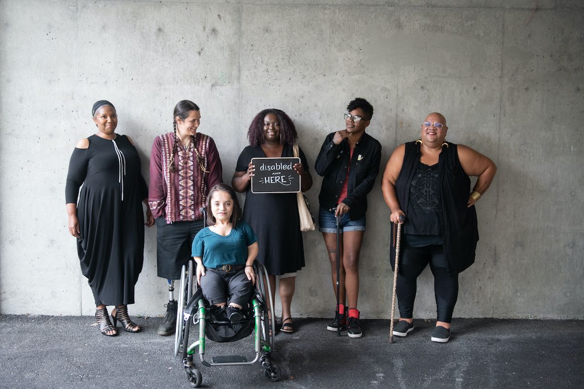 Six disabled people of color smile and pose in front of a concrete wall. Five people stand in the back, with the Black woman in the center holding up a chalkboard sign reading "disabled and here." A South Asian person in a wheelchair sits in front.